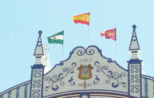 Las tres banderas lucen en el centro de la portada de la Feria de Abril desde esta mañana / Rubén Reina (@al_lawro)